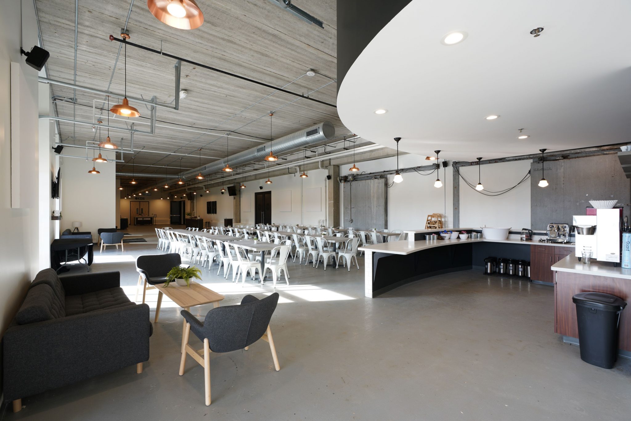 Industrial-modern cafeteria with exposed concrete ceilings, copper pendant lights, white cafe seating, and a lounge area.